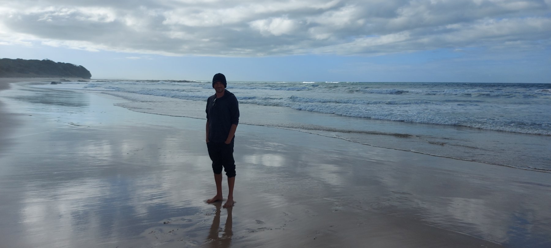 Nathaniel Calhoun, walking on a wide, wet beach with the surf and headland in the distance.